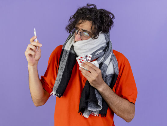 年轻人焦急的年轻白种人 戴着眼镜 戴着围巾 拿着体温计和药丸 看着隔离在紫色墙上的体温计男人疾病穿