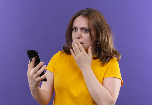 手持惊讶的年轻随意的女人手放在嘴上拿着手机在孤立的紫色墙上休闲电话手