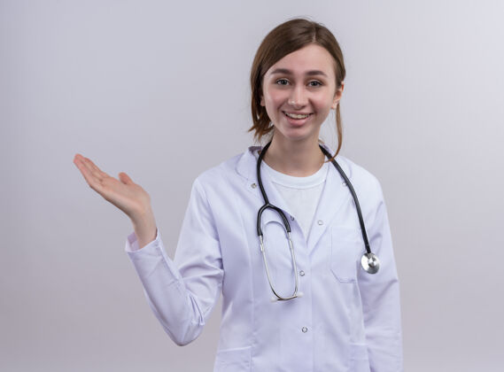 长袍面带微笑的年轻女医生 穿着医用长袍 戴着听诊器 在孤零零的白墙上露出空手年轻表演医生