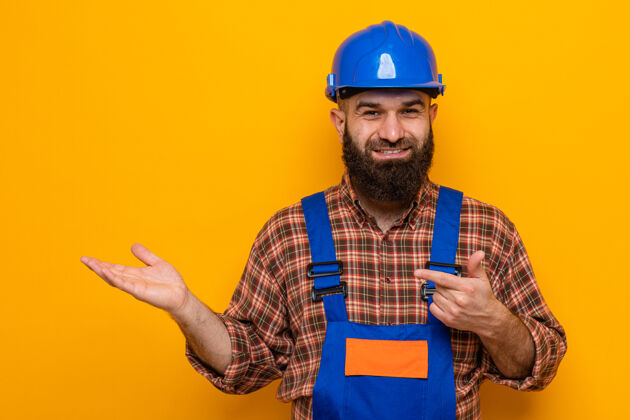 介绍留着胡须的建筑工人身着建筑制服 头戴安全帽 面带微笑 手把手用食指指向一边 愉快地展示着什么东西头盔手指手