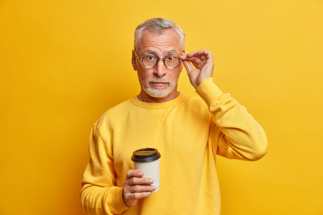拍摄惊讶的大胡子成熟男人手放在眼镜上喝外卖咖啡听到惊人的消息穿着休闲套头衫隔着鲜艳的黄色墙壁拿着反应老年人