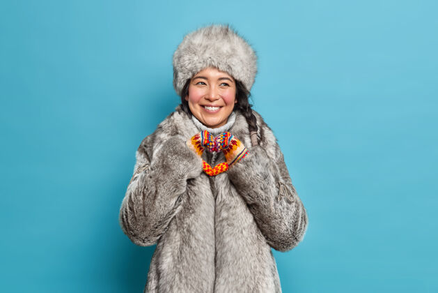 个人积极的斯堪的纳维亚女人在冬天外套做心形表示爱和关怀微笑愉快地有白色的牙齿隔着蓝色的墙北方黑发满意