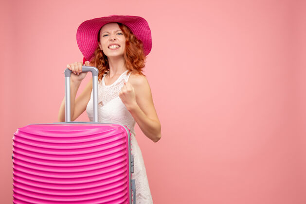 模特年轻的女性游客与粉红色的帽子和粉红色的墙壁上的包的正面视图度假旅行帽子
