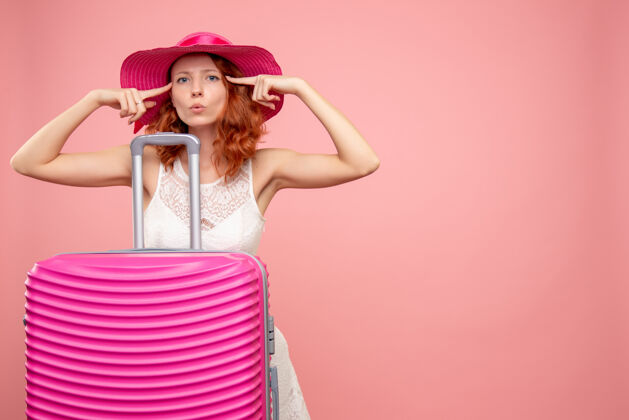 模特年轻的女性游客与粉红色的帽子和粉红色的墙壁上的包的正面视图肖像人度假