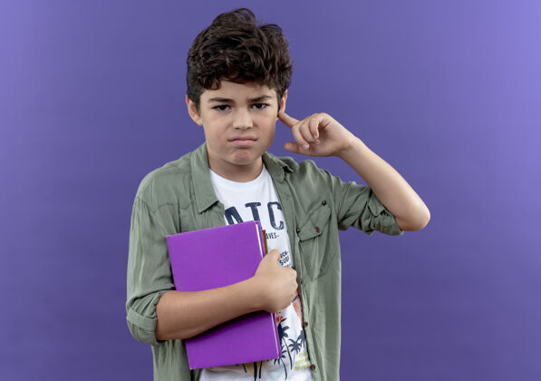 握着一个拿着书 把手指放在耳朵上的小男孩被隔离在紫色的墙上讨厌小小学生