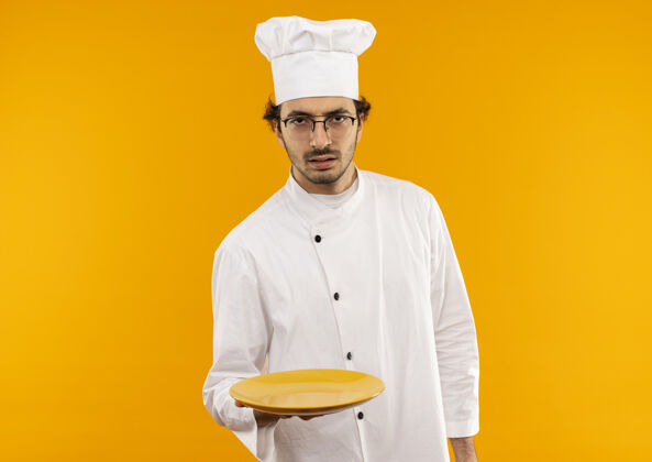 严格严格年轻的男厨师穿着厨师制服 戴着眼镜把盘子隔离在黄色的墙上厨师年轻穿着