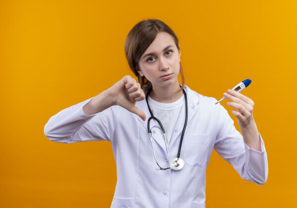 长袍年轻的女医生穿着医用长袍 手持听诊器 拿着温度计 拇指朝下放在隔离的橙色墙上年轻人展示放下