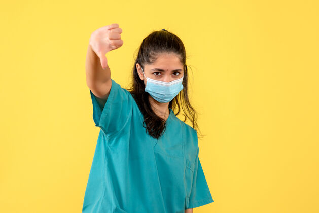 医学黄色墙上戴着医用口罩的漂亮女医生大拇指朝下标志的正面图向下微笑脸
