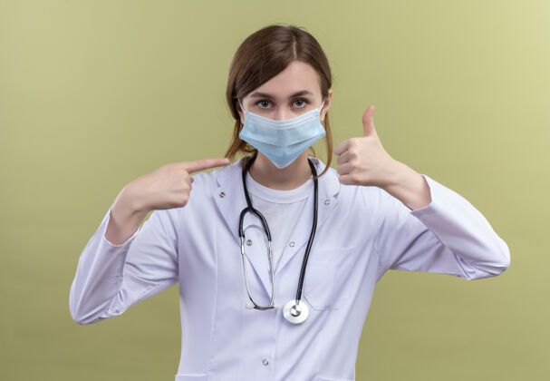 医生年轻的女医生穿着医用长袍 戴着口罩和听诊器 在隔离的绿色墙上竖起大拇指指着它穿长袍指尖