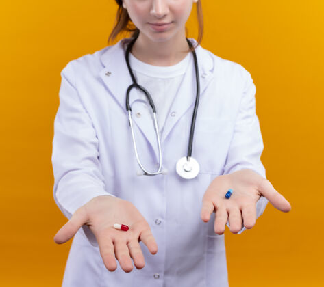 伸展年轻的女医生穿着医用长袍和听诊器在隔离的橙色墙上伸展医用胶囊医生女长袍