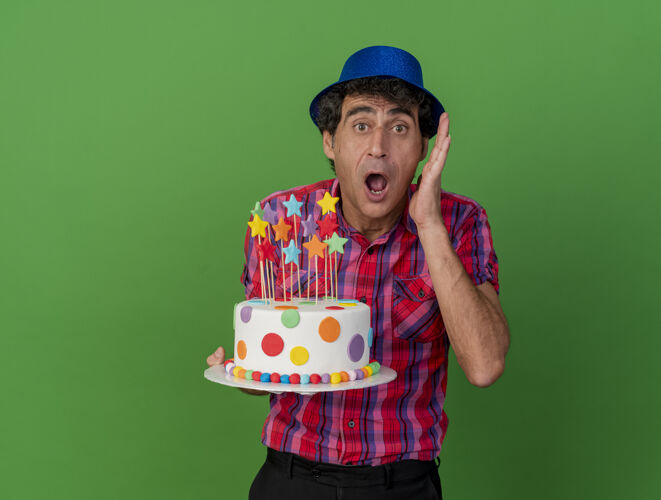 蛋糕震惊的中年白种人党男子戴着党的帽子拿着生日蛋糕看着相机保持手靠近头部看着相机隔离在绿色背景与复制空间聚会头手