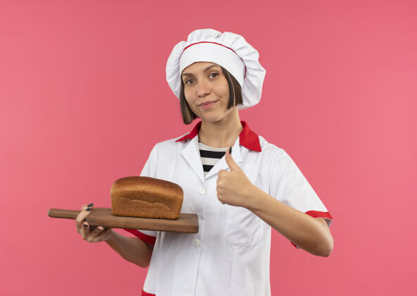 面包自信的年轻女厨师身着厨师制服 手拿着放着面包的砧板 在粉红色的地板上孤立地竖起大拇指女向上举行