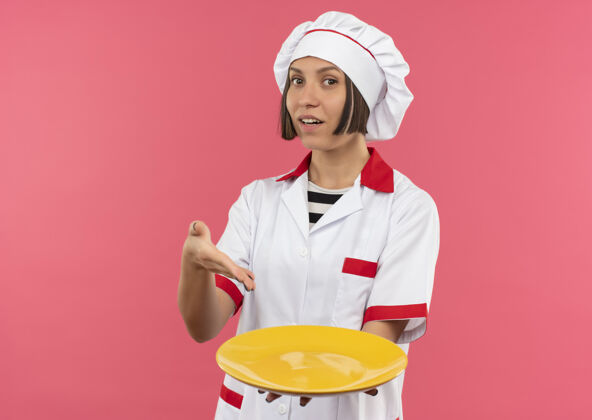 复制印象深刻的年轻女厨师在厨师制服举行 并指着空盘子孤立的粉红色与复制空间制服空间持有