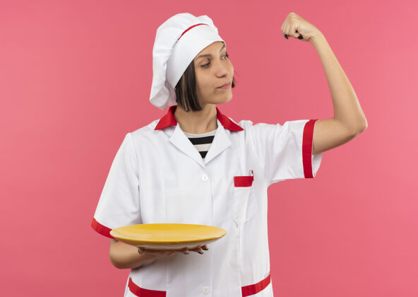 年轻自信的年轻女厨师身着厨师制服 指着结实的托板 看着孤立在粉色地板上的手臂盘子粉色厨师