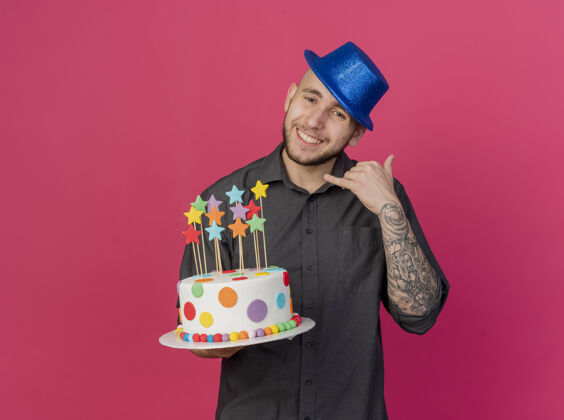 蛋糕微笑的年轻帅哥斯拉夫党人戴着党帽拿着生日蛋糕与明星们看着相机做通话手势在深红色的背景与复制空间隔离电话星星相机