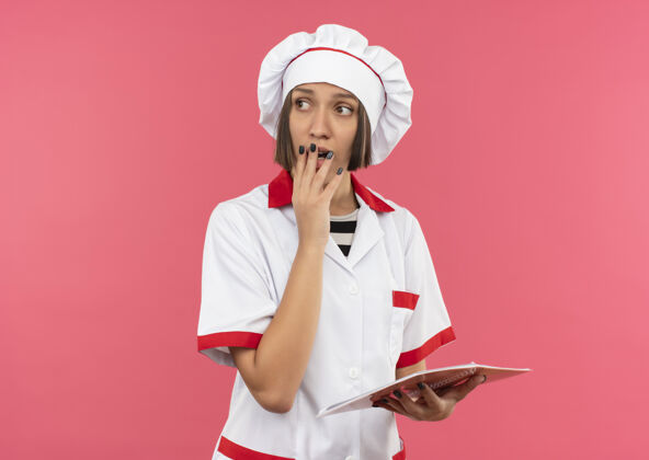 印象印象深刻的年轻女厨师穿着厨师制服拿着便笺簿把手放在嘴上 看着粉红色的一面隔离着复印空间笔记粉红厨师