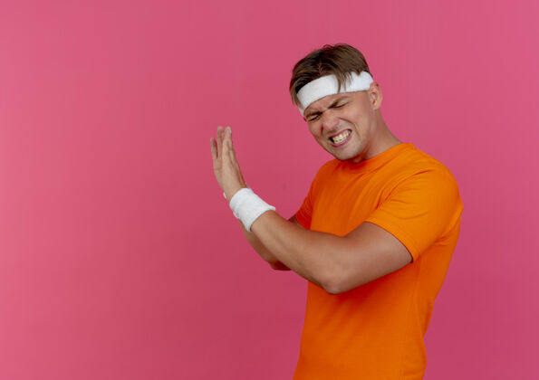 佩戴恼怒的年轻英俊的运动型男子戴着头带和腕带站在侧视图中表示不 闭着眼睛孤立在粉红色的复制空间眼睛帅气运动