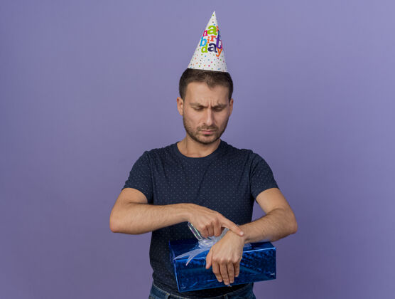 帅气戴着生日帽的白种人帅哥拿着礼物盒 看着紫色背景上的礼物盒 盒子上留着复印件帽子礼物拿着