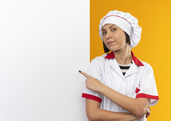 站立自信的年轻女厨师身着厨师制服站在白色的墙壁前 指着隔离在橙色的墙壁 留有复印空间前面信心指向