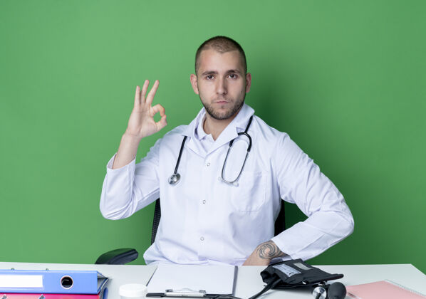 男性自信的年轻男性医生穿着医用长袍和听诊器坐在办公桌旁 带着工作工具 在绿色的隔离标志上做得很好好的绿色标志