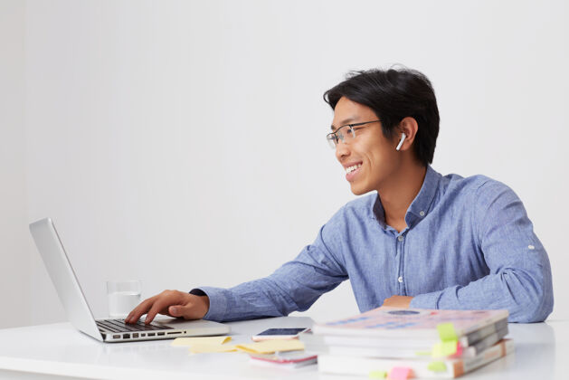 亚洲人戴着眼镜 穿着蓝色衬衫 微笑着的亚洲年轻商人 在白墙上使用无线耳机和笔记本电脑进行视频通信聊天男人打字