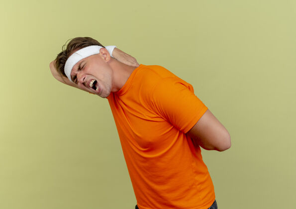 视野疼痛的年轻英俊的运动型男子戴着头带和腕带站在侧面图把双手放在背上隔离在橄榄绿站立运动年轻