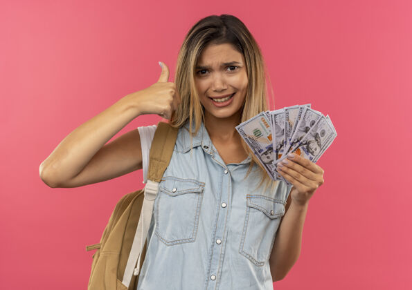 女孩自信的年轻漂亮的女学生 背着包 手里拿着钱 在粉红色的地板上孤立地竖起大拇指拇指持有背部