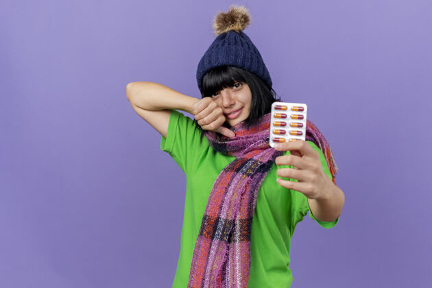 包年轻的白种女孩戴着冬天的帽子 戴着围巾 伸出一包胶囊 大拇指朝上 看着紫色背景上的相机 还有复制空间围巾请伸展