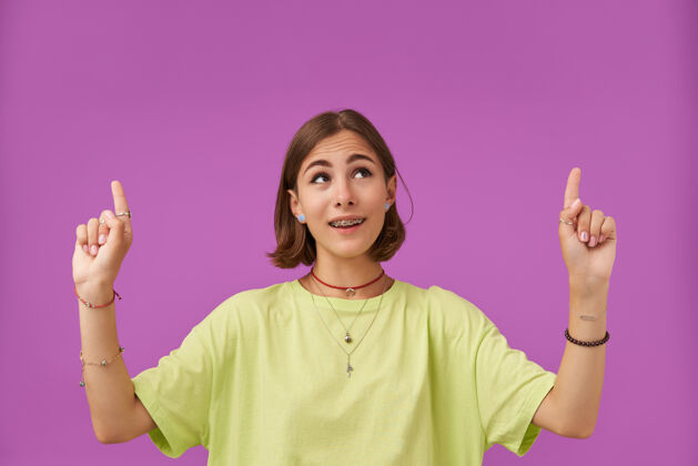 服装迷人的 漂亮的 神奇女孩的肖像手指指向紫色墙上的复制空间穿着绿色t恤 牙套 项链 手镯和戒指短发面部T恤