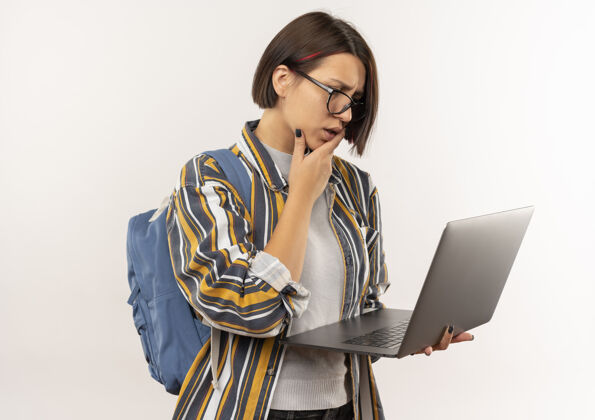 手困惑的年轻女学生戴着眼镜 背着书包拿着笔记本电脑 手放在下巴上 孤立地看着白色穿背笔记本电脑