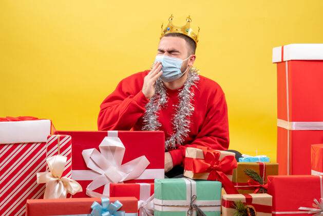 坐着正面图：坐在圣诞节周围的年轻男性戴着黄色背景的面具出现欢乐病毒大流行