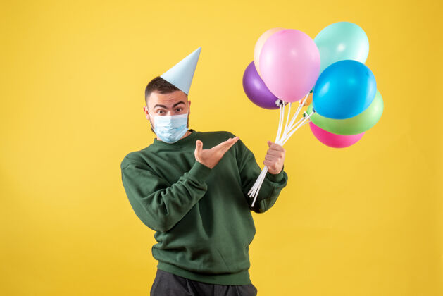 病毒正面图：黄色背景上戴着面具手持彩色气球的年轻男性庆祝人面具