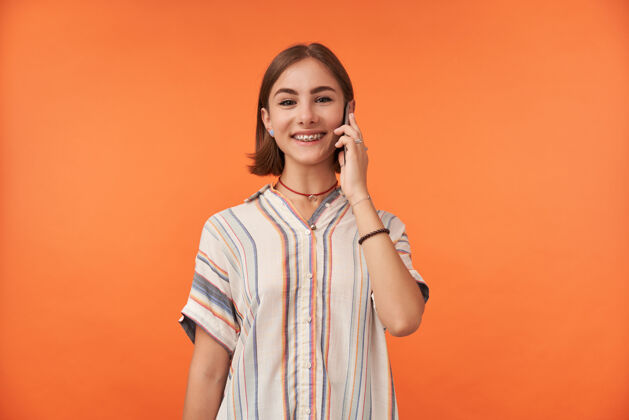 成人一个迷人的年轻女子微笑的肖像在智能手机上交谈 穿着条纹衬衫 戴着牙套和手镯站在橙色的墙上孤立衬衫年轻微笑