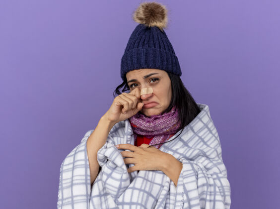 包裹悲伤的年轻白种人生病的女孩戴着冬天的帽子 围着方格布围巾 看着鼻子上贴着医用膏药的相机 在紫色背景上擦眼泪相机悲伤医疗