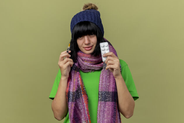 围巾虚弱的年轻病高加索女孩戴着冬天的帽子和围巾拿着注射器和一包药片看着相机隔离在橄榄绿的背景与复制空间医疗虚弱年轻