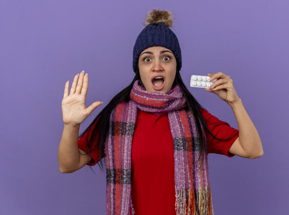 年轻印象深刻的年轻白种人生病的女孩戴着冬天的帽子和围巾显示包平板电脑看着相机显示空手孤立在紫色的背景围巾空相机