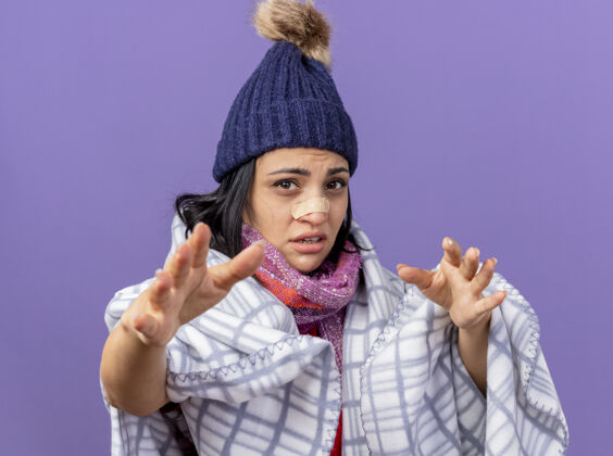 石膏虚弱的年轻白种人生病的女孩戴着冬天的帽子 围巾裹着格子布 看着鼻子上贴着医用膏药的相机 向隔离在紫色背景上的相机伸出双手疾病虚弱帽子