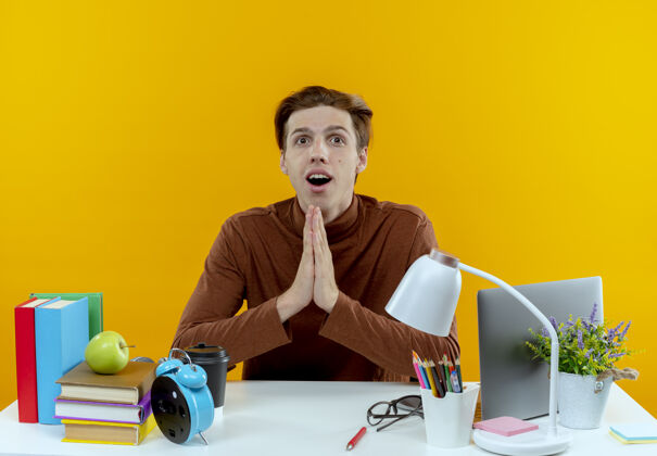 书桌惊讶的年轻学生男孩坐在书桌旁 黄色的墙壁上展示着祈祷的手势坐着工具惊喜