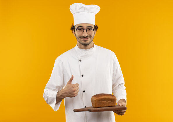眼镜穿着厨师制服 戴着眼镜 拿着面包在砧板上的年轻男厨师微笑着 他的大拇指孤立地竖立在黄色的墙上面包切割烹饪