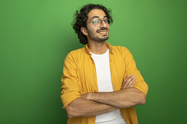 帅气微笑着的年轻英俊的白人男子戴着眼镜站着 以封闭的姿势看着隔离在绿色背景上的相机 还有复印空间微笑封闭姿势