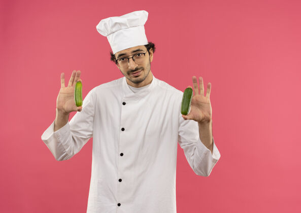眼镜年轻的男厨师穿着厨师制服 戴着眼镜 把黄瓜隔离在粉红色的墙上男请戴着
