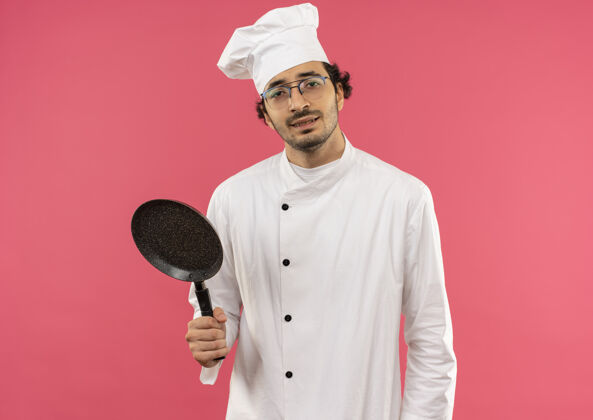 男年轻的男厨师穿着厨师制服 戴着眼镜 拿着平底锅孤立地站在粉红色的墙上平底锅年轻烹饪
