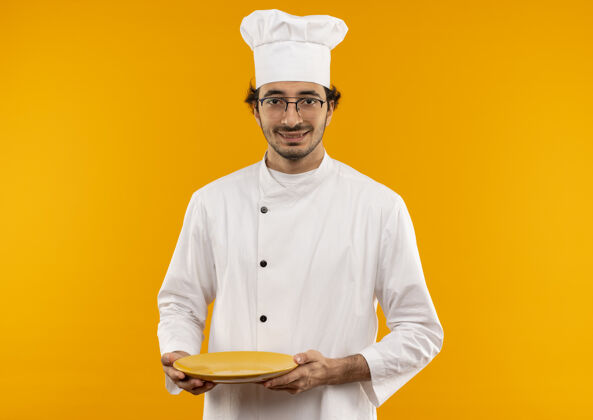 盘子面带微笑的年轻男厨师穿着厨师制服 戴着眼镜 把盘子隔离在黄色的墙上制服穿拿着