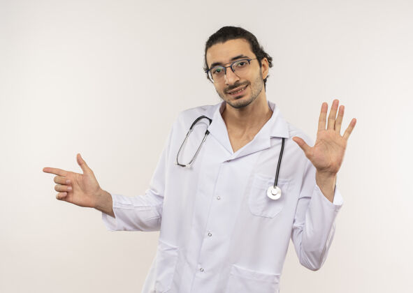 听诊器年轻的男医生戴着眼镜 身穿白色长袍 听诊器显示出停止的姿势 并指着隔离的白色墙壁上有复印空间的一侧眼镜显示请