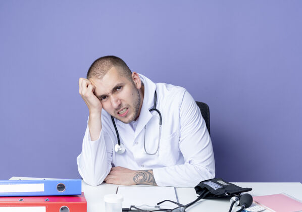 穿恼怒的年轻男医生穿着医用长袍和听诊器坐在办公桌前 手放在头上 脸上一片紫色手听诊器紫色