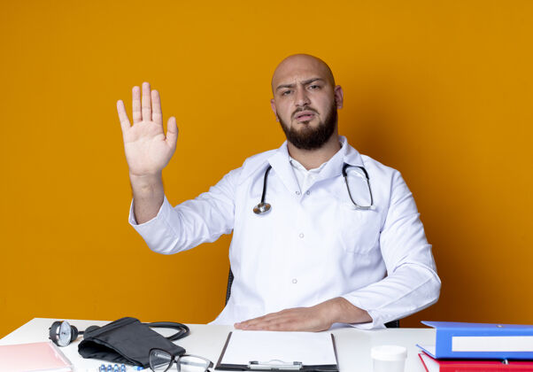 穿严格年轻的秃头男医生穿着医用长袍和听诊器坐在工作台上 医疗工具显示停止手势隔离在橙色的墙上显示听诊器医生