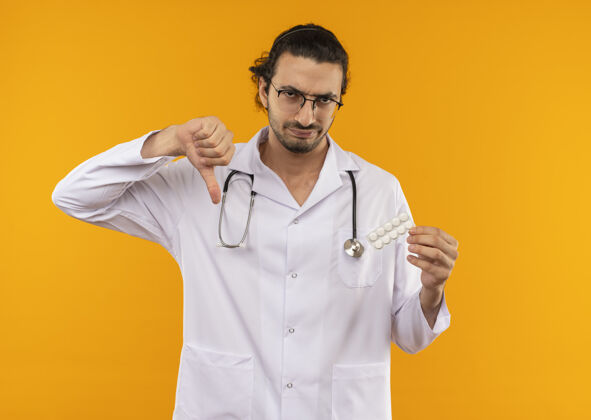 年轻戴着医用眼镜 穿着医用长袍 手持听诊器 拿着药片的悲伤的年轻医生 他的大拇指放在隔离的黄色墙壁上 还有复印空间眼镜穿听诊器