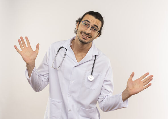 男性微笑着的年轻男医生戴着眼镜 穿着白色长袍 带着听诊器 在隔离的白色墙壁上摊开双手 留着复印空间手眼镜穿