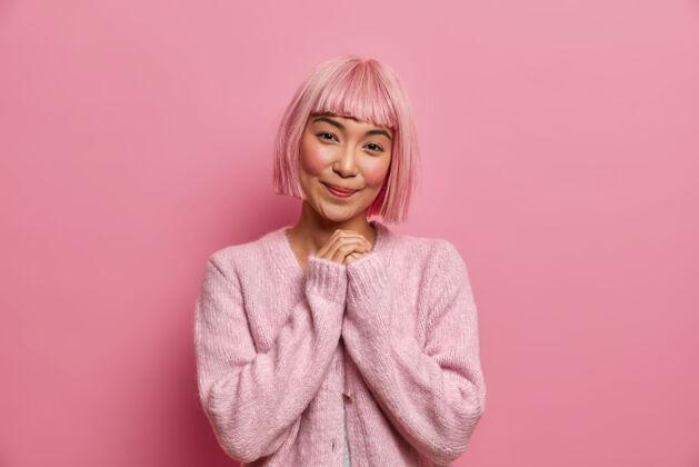 发型美丽的亚洲女人的照片 心情很好 双手合十 有迷人的笑脸 想着什么好笑的穿粉红色的套头衫站在室内假发手内容