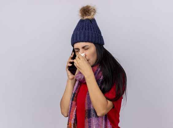 冬天皱眉的年轻白种人生病的女孩戴着冬天的帽子和围巾站在侧视图讲电话擦鼻子用餐巾隔离在白色背景与复印空间餐巾白色疾病